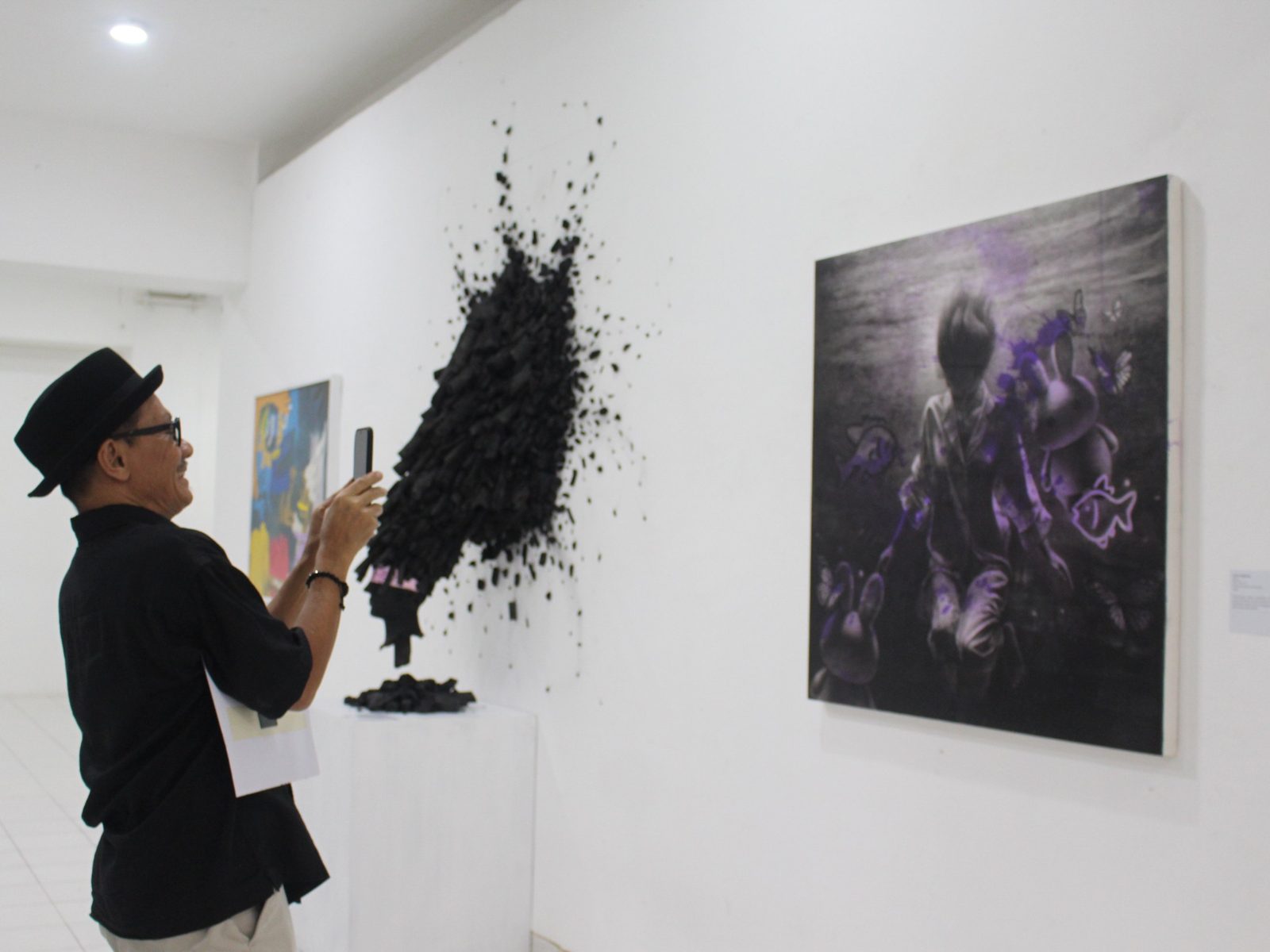 Sunyi hanging on a white gallery wall beside a black charcoal sculpture, with a visitor photographing the work